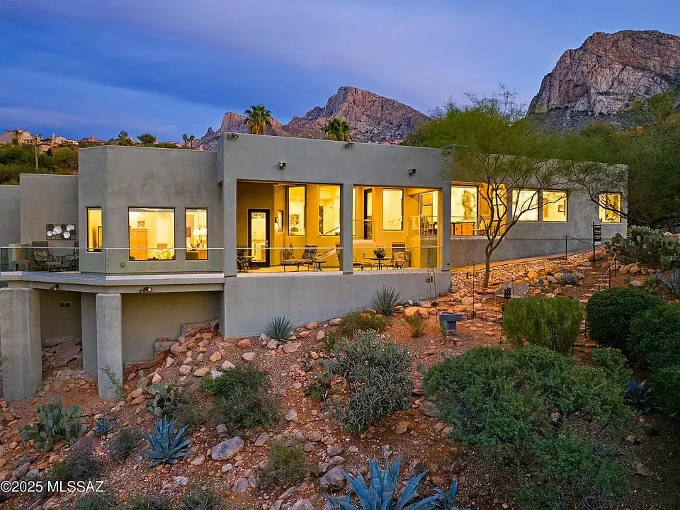 Modern house with large windows, surrounded by desert landscape and mountains at dusk.