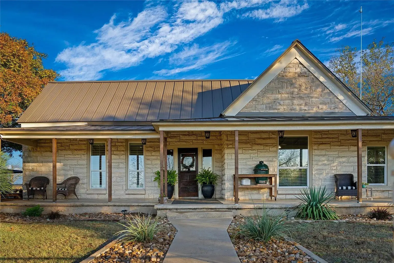 Single-story stone house with a metal roof, front porch, and chairs, set against a blue sky.