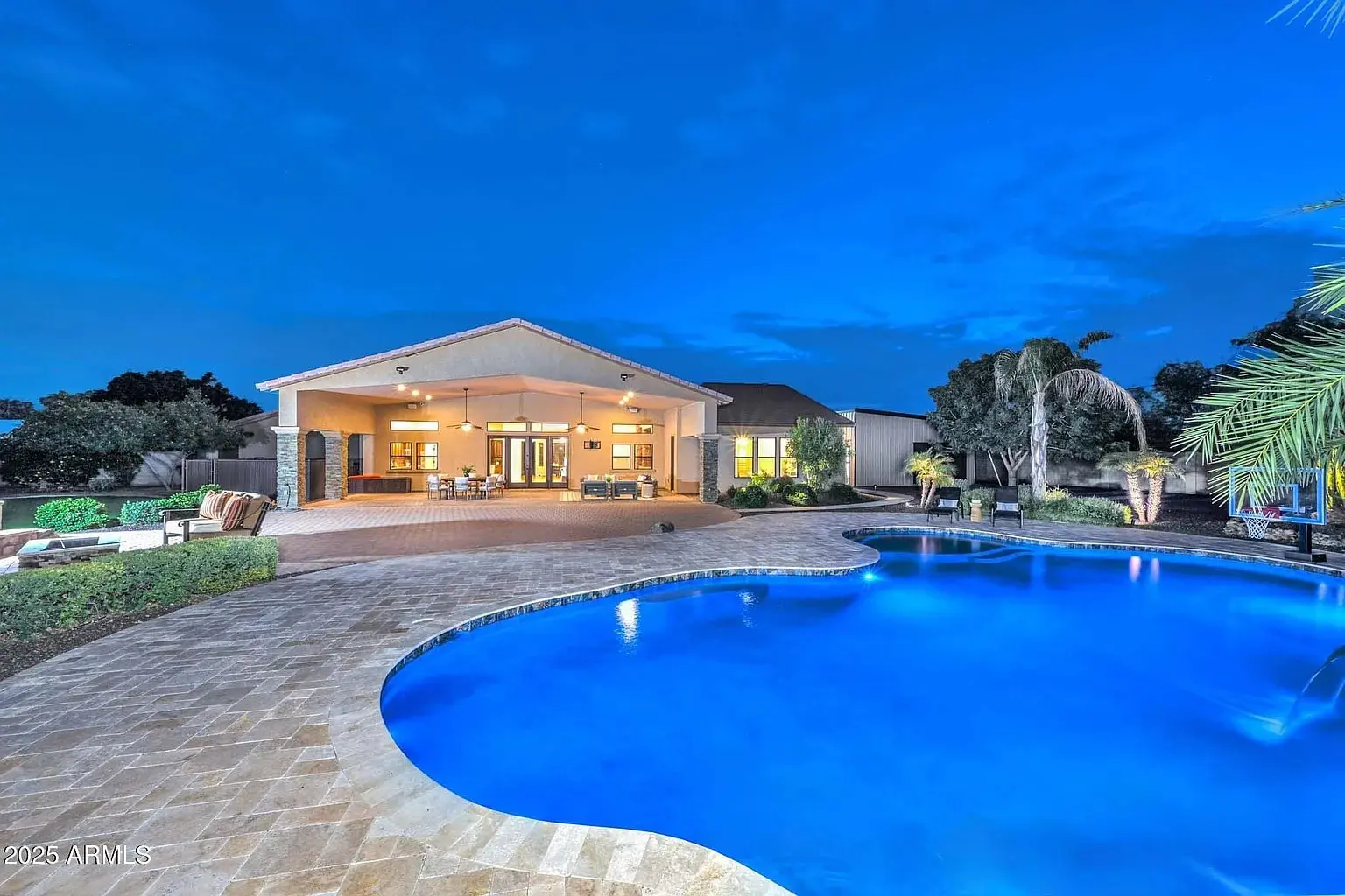 Spacious backyard with illuminated pool, patio seating, and a house under a twilight sky.