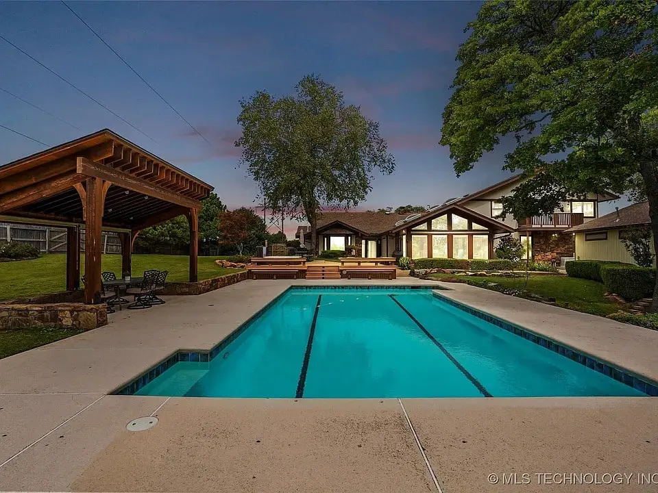 Outdoor pool area with a covered seating space, surrounded by trees and a modern house.