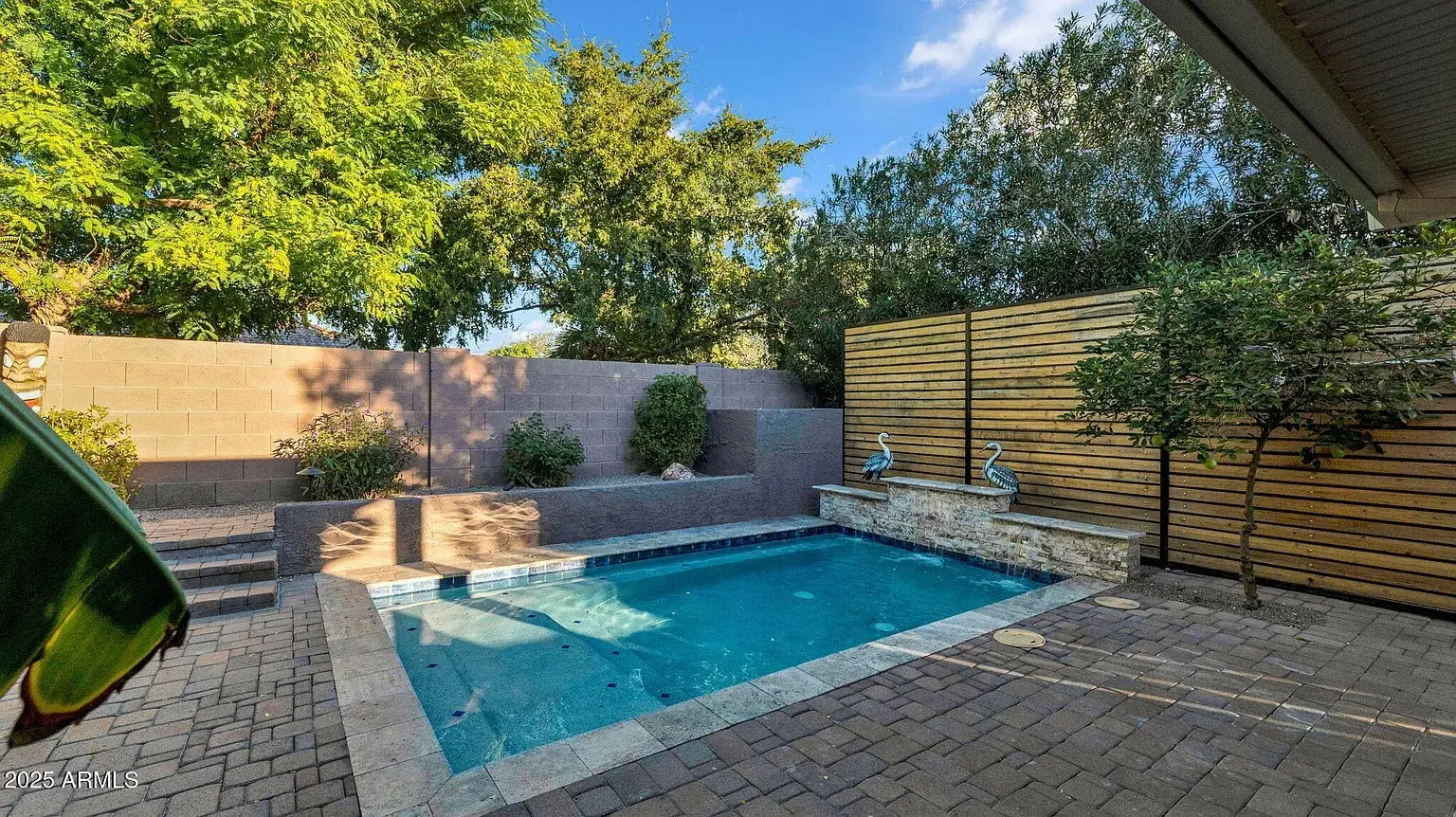 Small backyard pool with stone and wood fencing, surrounded by trees and paved patio area.