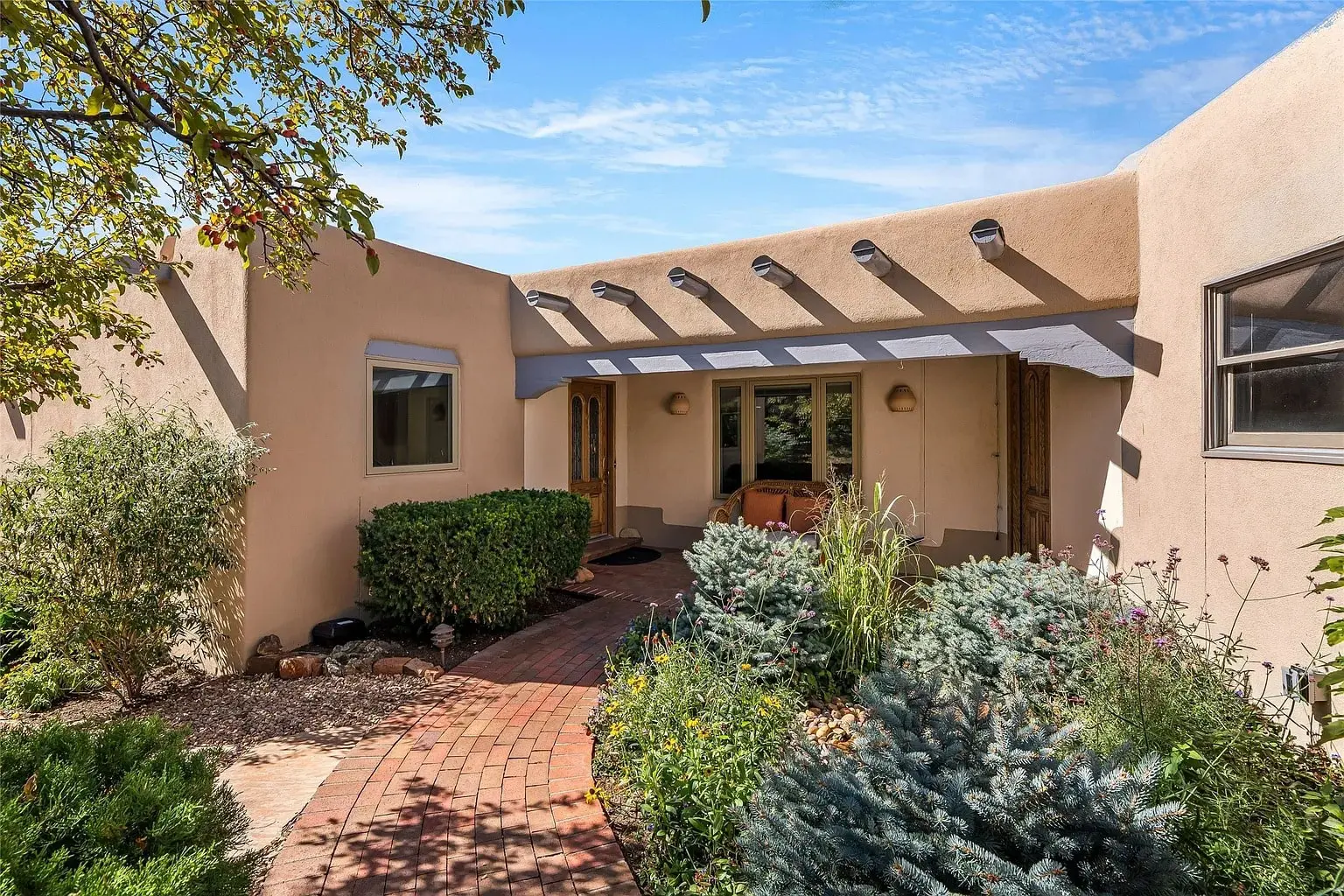 Modern adobe-style house entrance with a brick pathway, greenery, and a wooden pergola.