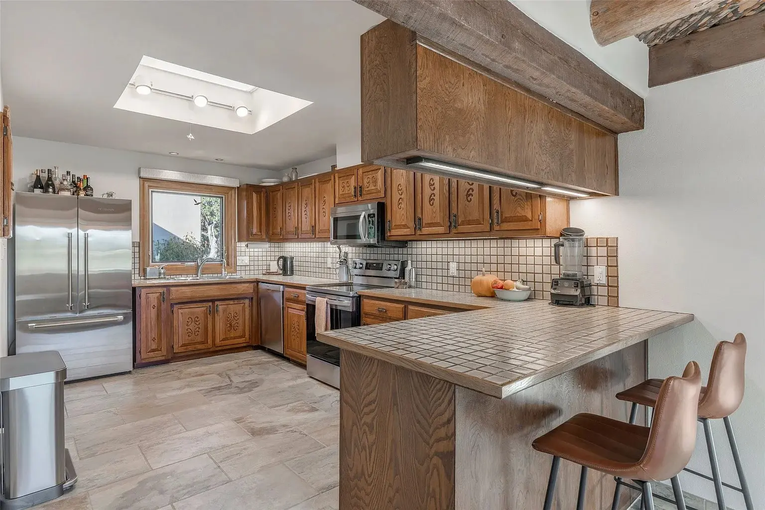 Modern kitchen with wooden cabinets, stainless steel appliances, tiled countertops, and bar seating.