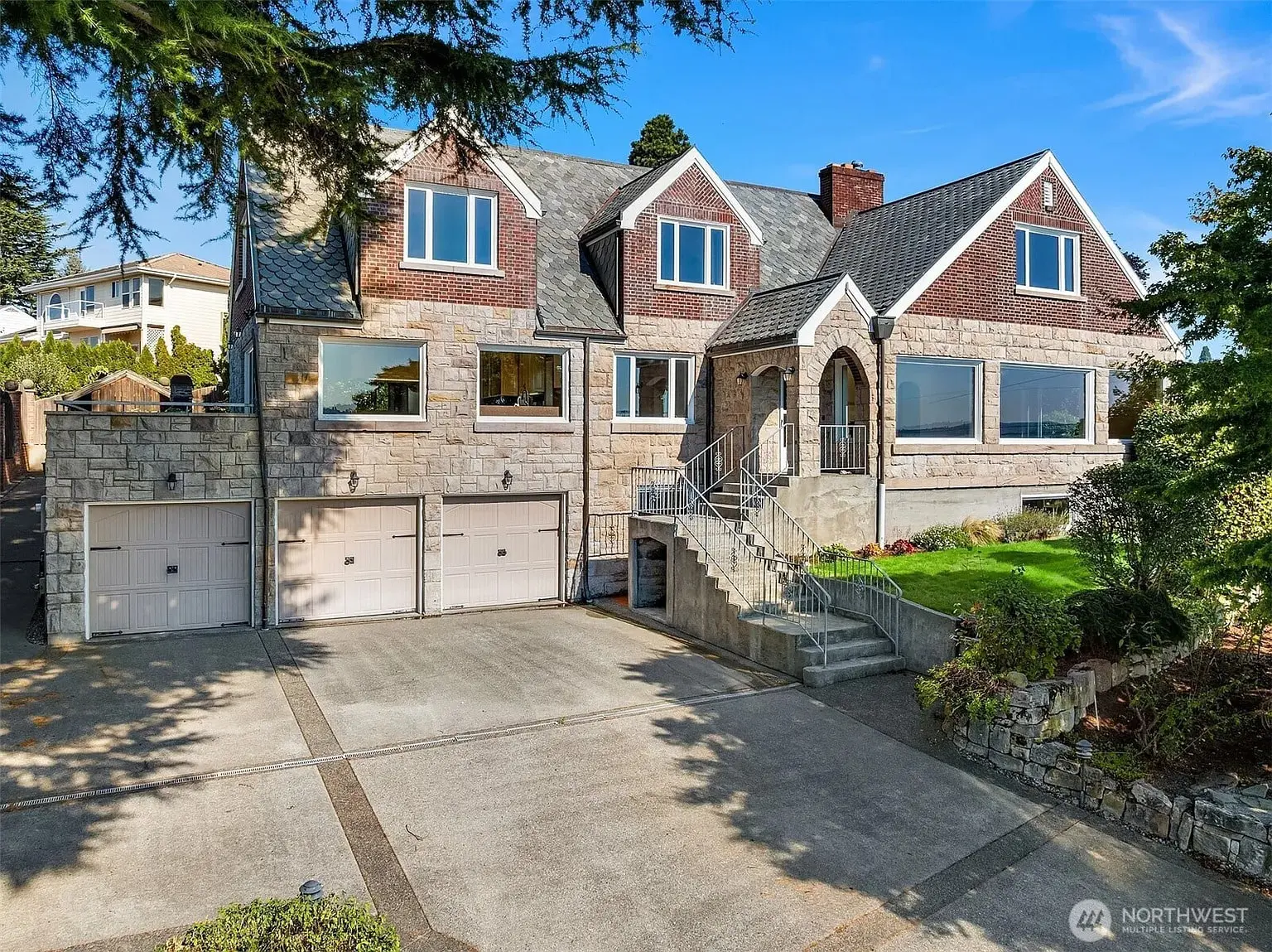 Large stone house with three garage doors, stairs leading to entrance, and a well-maintained lawn.