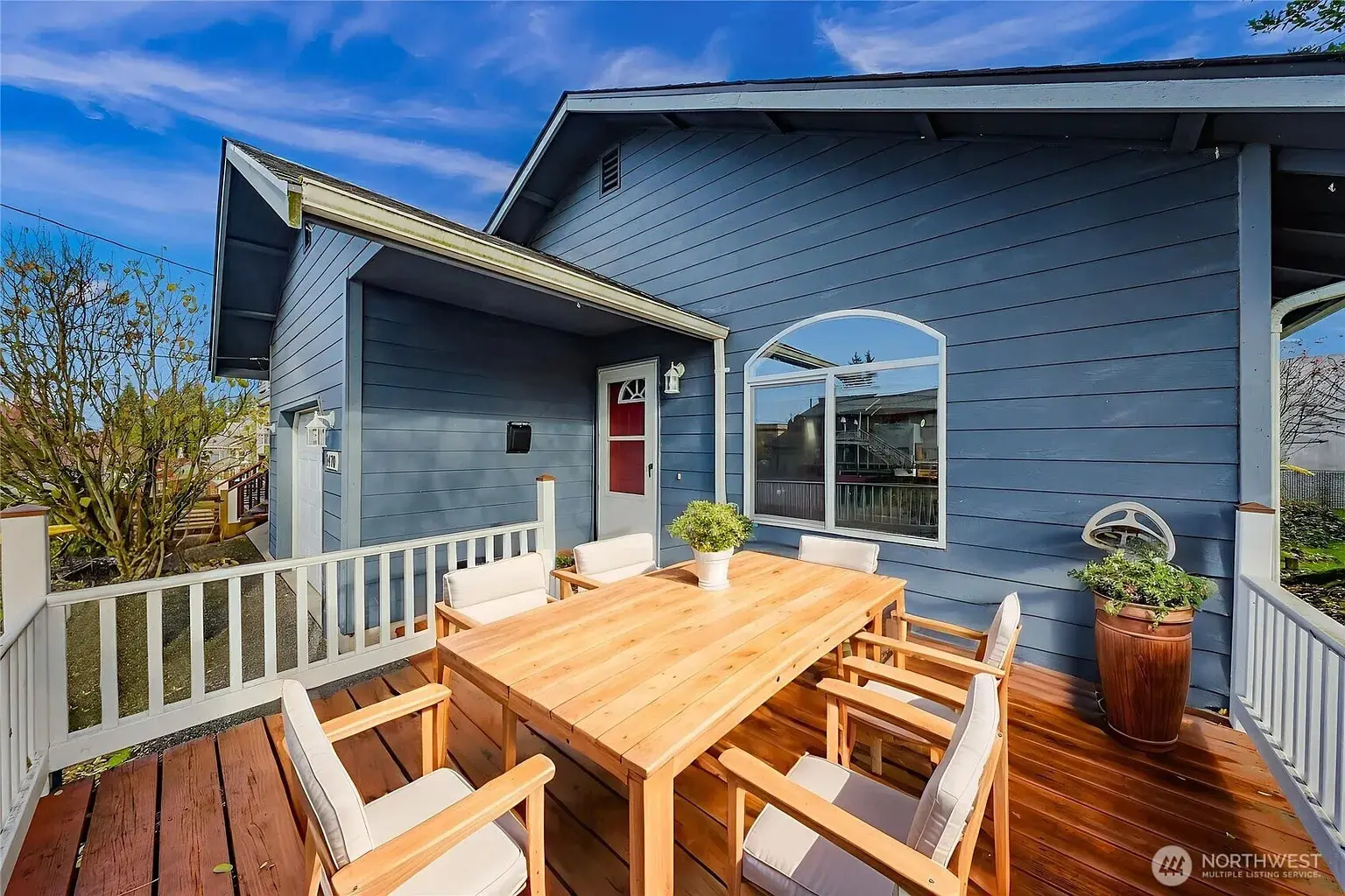 Wooden deck with outdoor dining set and potted plants, attached to a blue house under a clear sky.