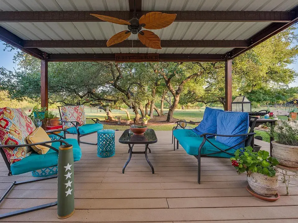 Covered patio with colorful cushioned chairs, table, plants, and a ceiling fan, overlooking a garden.