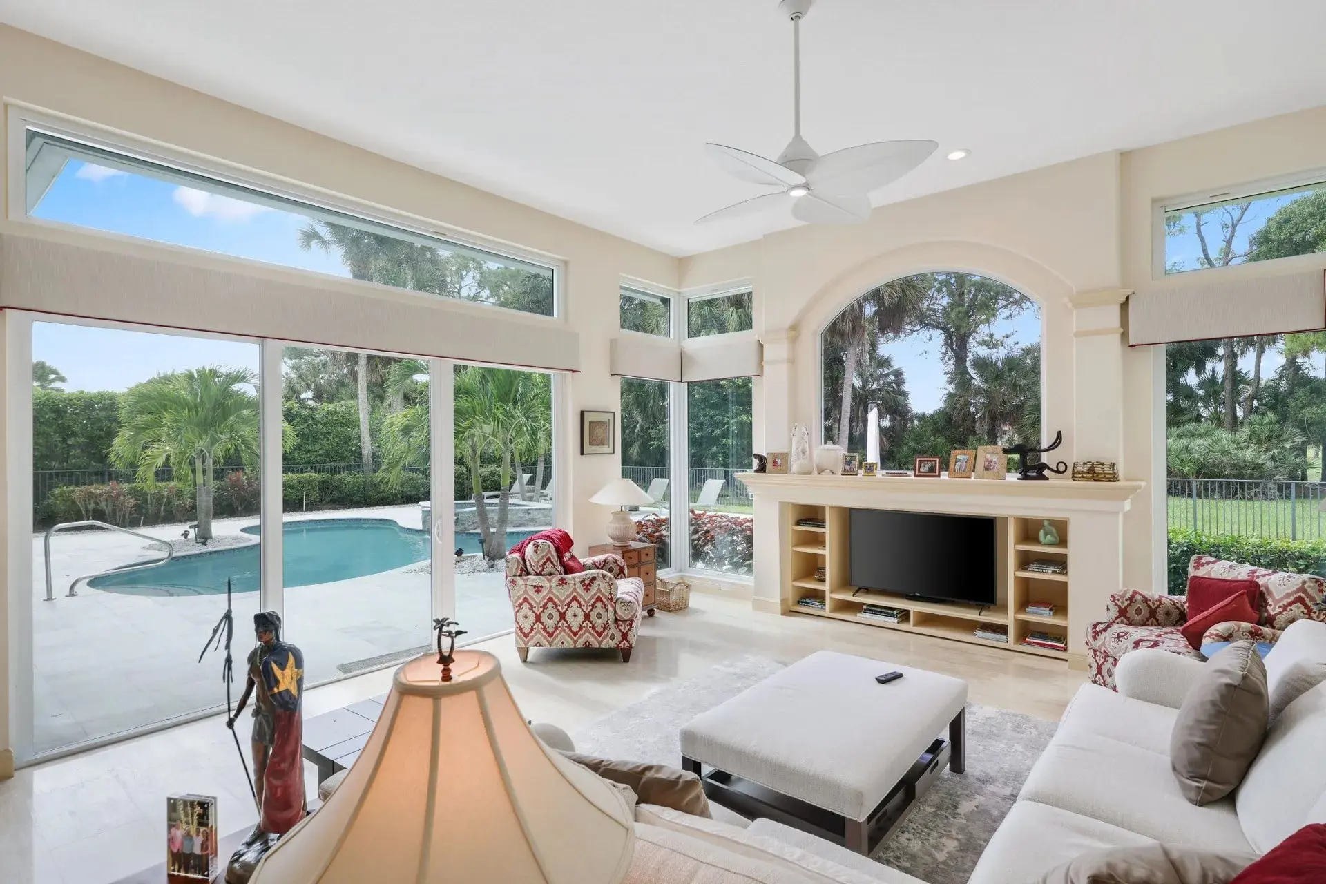 Spacious living room with large windows, pool view, modern furniture, and a ceiling fan.