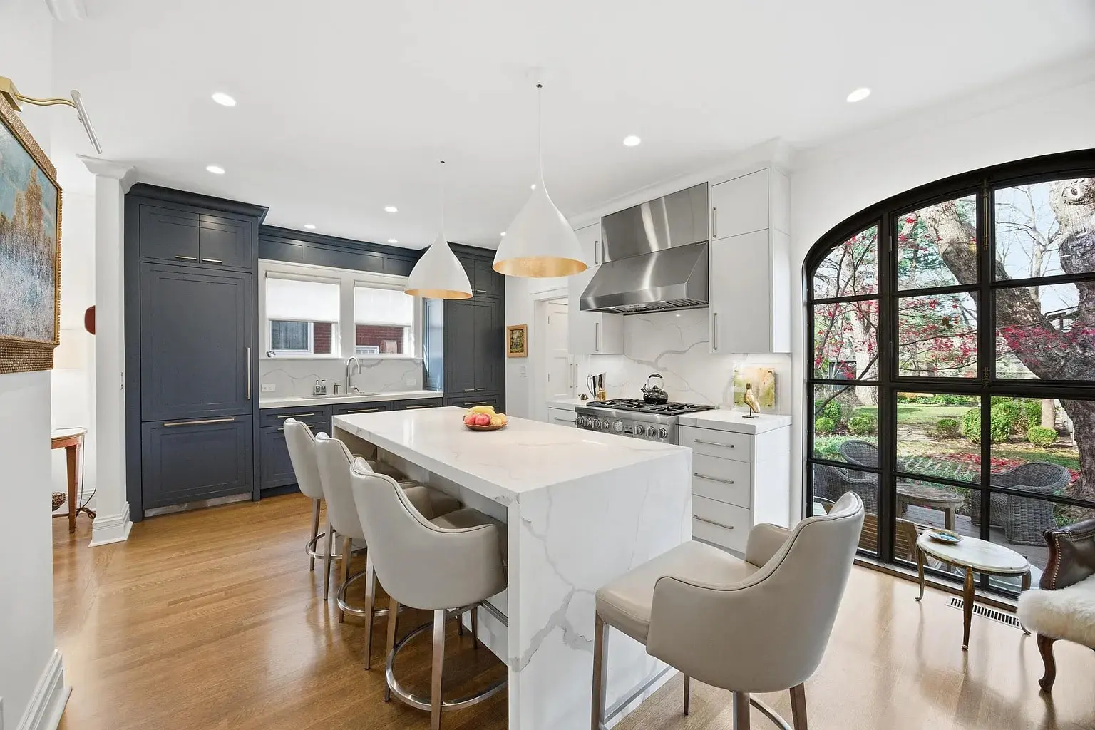 Modern kitchen with marble island, six chairs, dark cabinets, and large arched window overlooking garden.