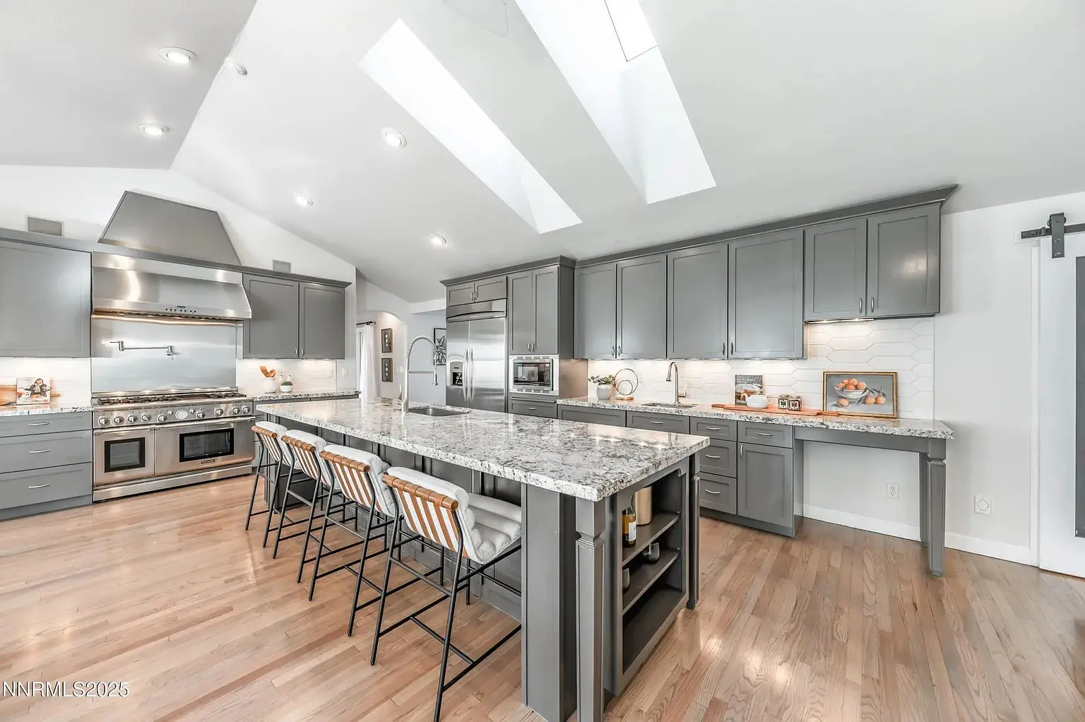 Modern kitchen with gray cabinets, large island with barstools, stainless steel appliances, and wooden flooring.