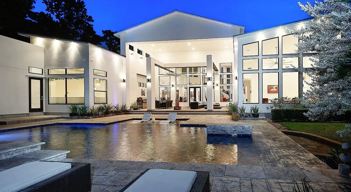 Modern house with large windows, illuminated at night, featuring a pool and patio area.