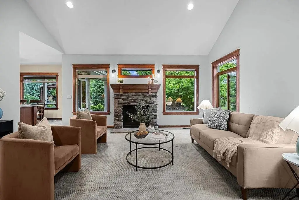 Living room with beige sofa, two brown chairs, stone fireplace, and large windows overlooking greenery.