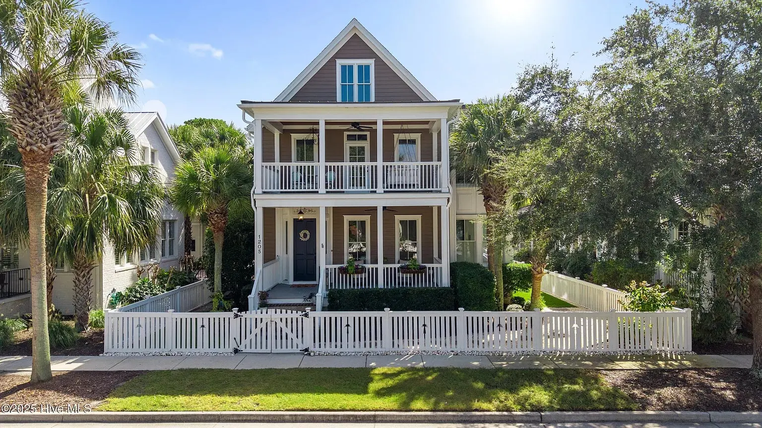 Two-story house with a front porch, white fence, and surrounding palm trees.