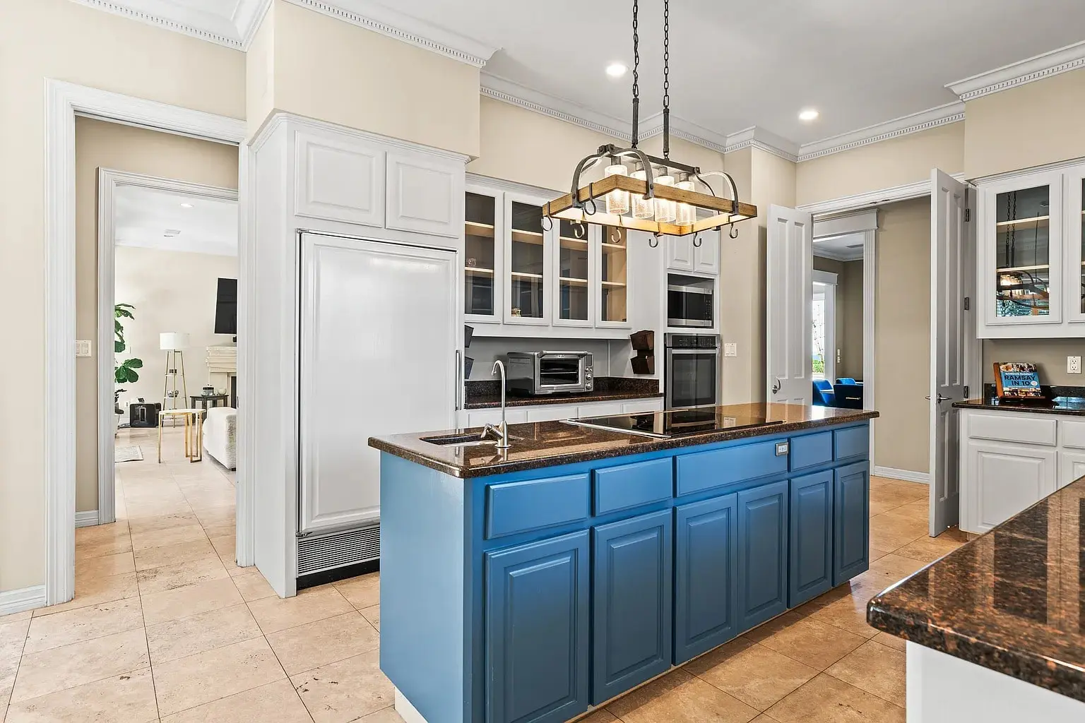 Modern kitchen with blue island, white cabinets, and hanging light fixture. Open doors lead to living area.
