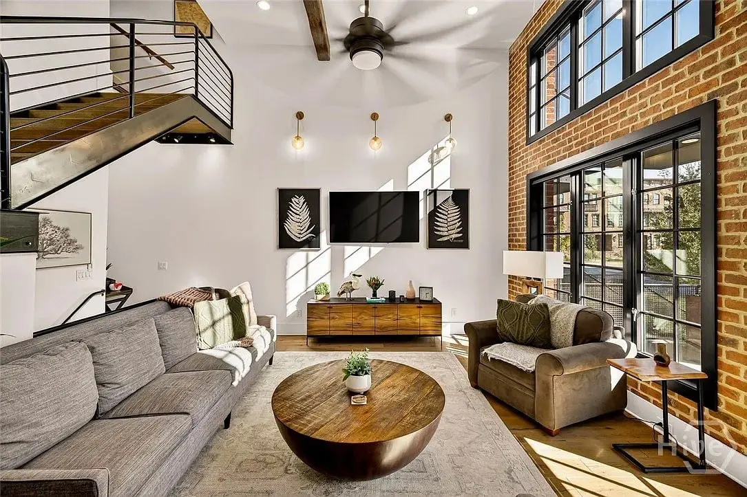 Modern living room with brick wall, large windows, gray sofa, armchair, and wooden coffee table.