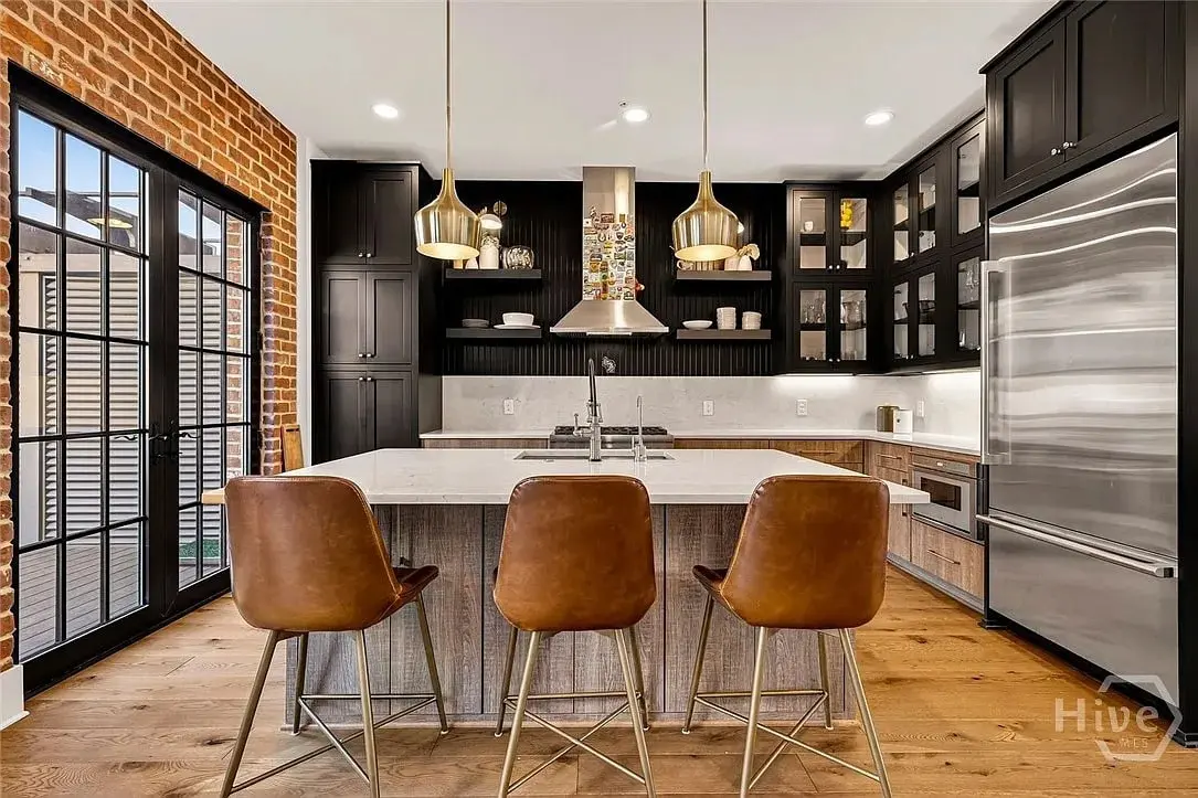 Modern kitchen with brick wall, large island, brown leather stools, and stainless steel appliances.