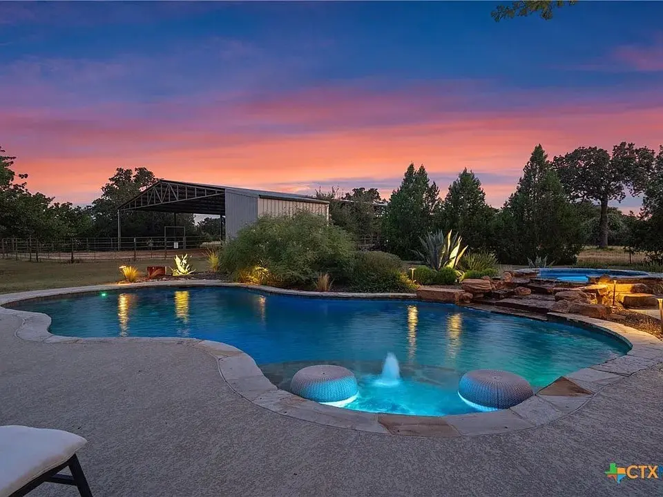 Backyard pool at sunset with trees, a barn, and colorful sky in the background.