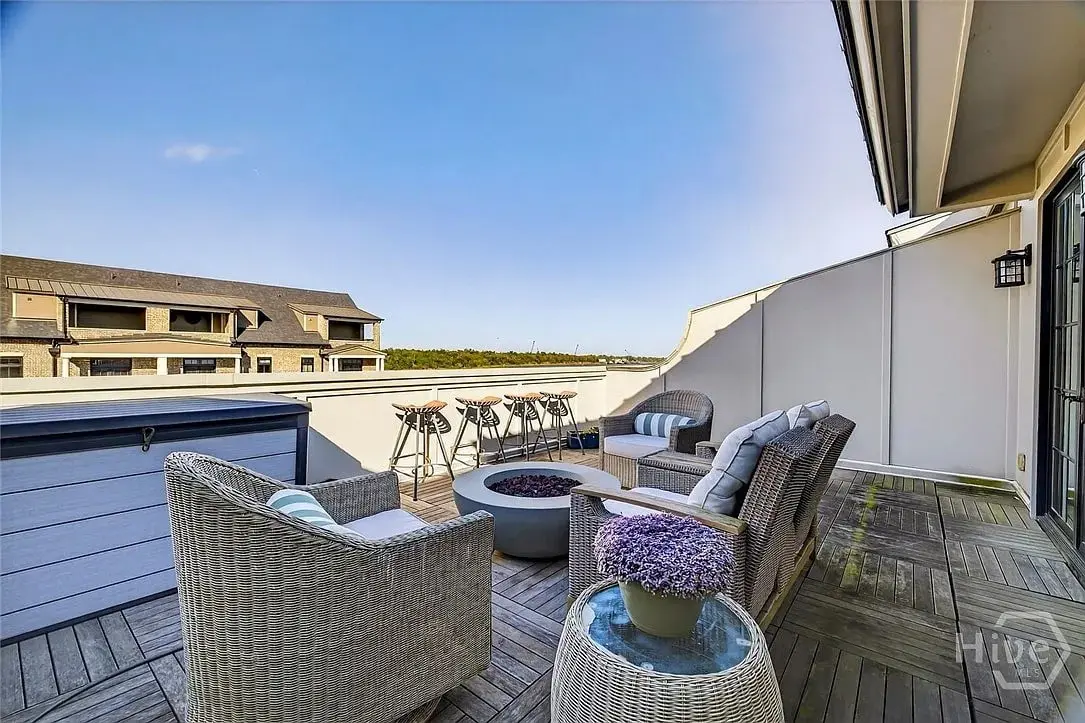 Rooftop patio with wicker chairs, a fire pit, potted flowers, and a distant view of buildings.