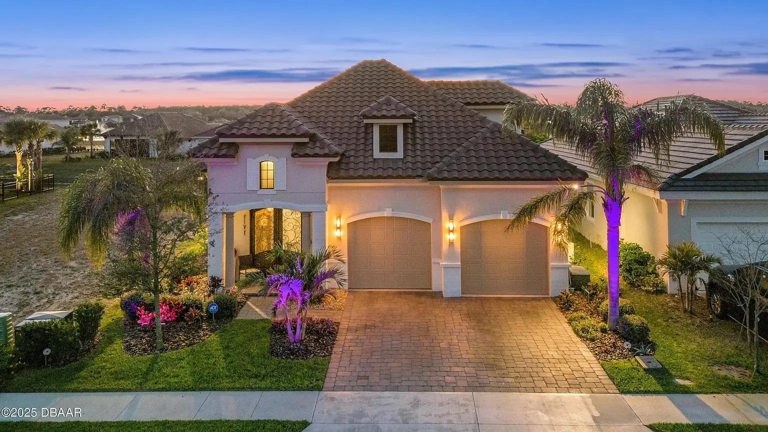 A Mediterranean-style house with a tiled roof, lit by exterior lights at sunset.