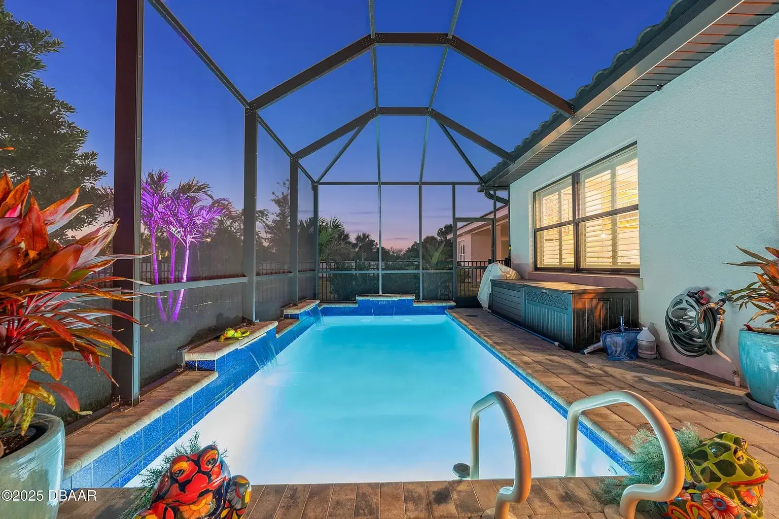 Enclosed backyard pool at dusk with colorful lighting and surrounding plants.