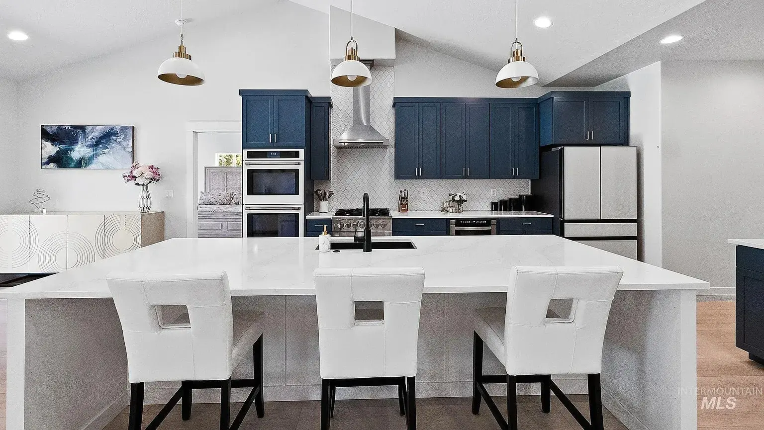 Modern kitchen with blue cabinets, white countertops, stainless steel appliances, and three white chairs at the island.