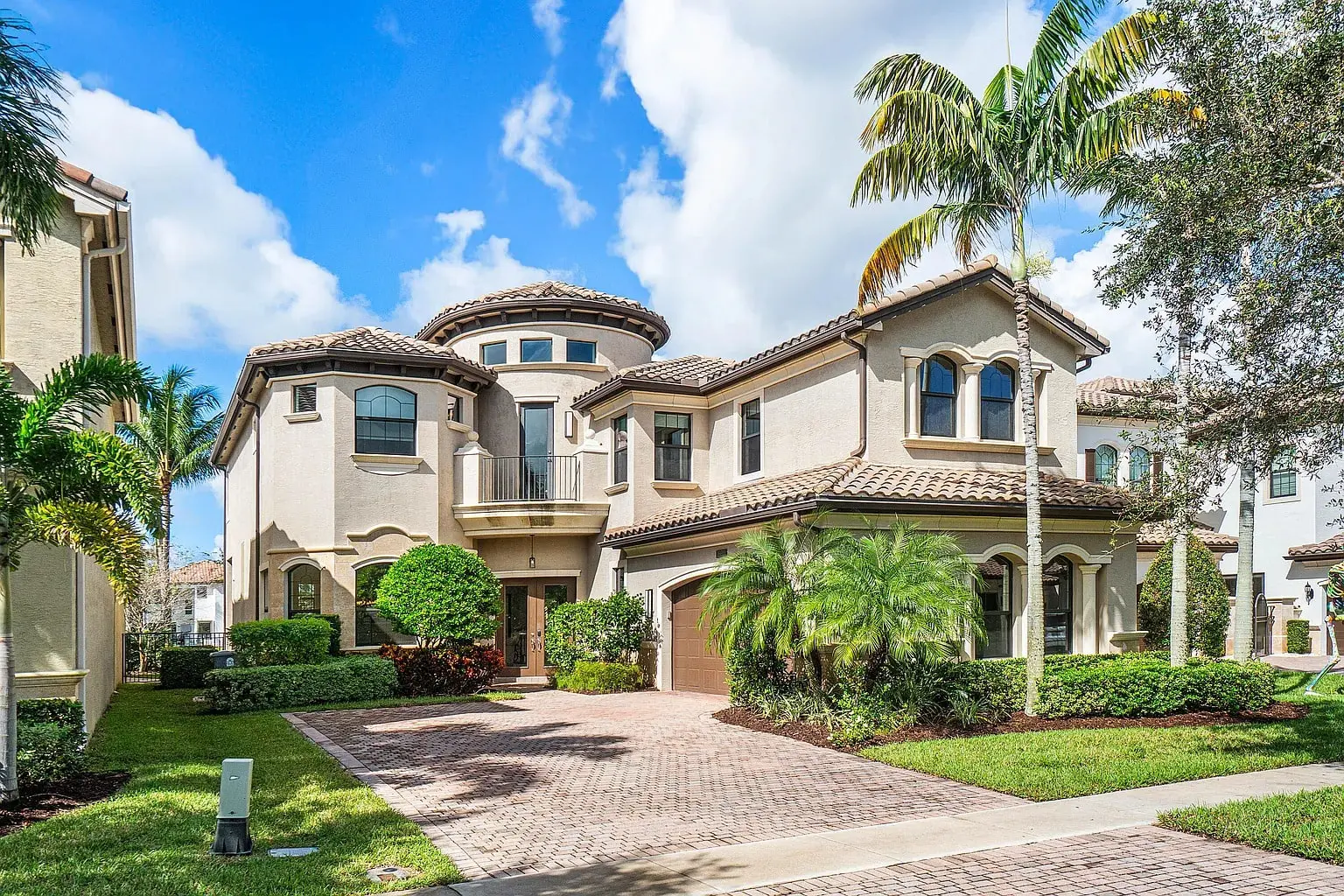 Large Mediterranean-style house with a tiled roof, palm trees, and a brick driveway under a blue sky.