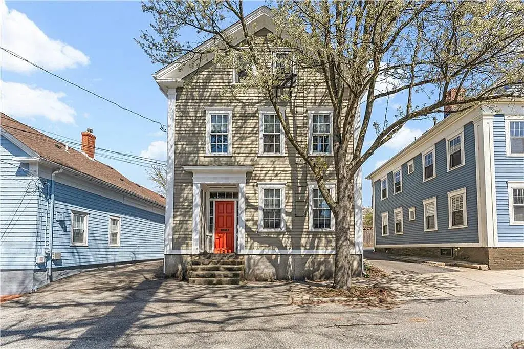 Historic two-story house with a red door, flanked by blue houses, and a tree in front.