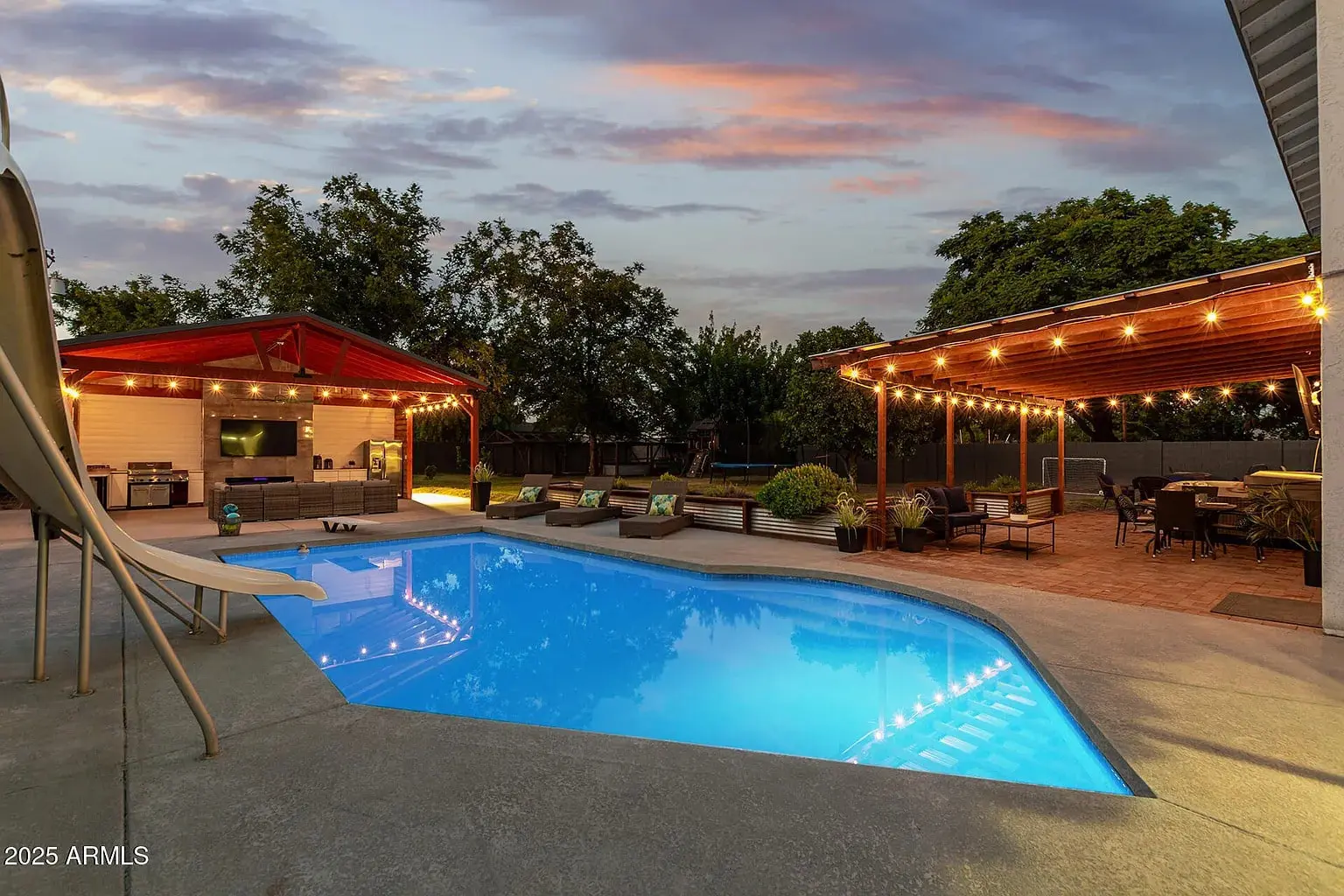 Backyard with illuminated pool, patio, string lights, outdoor kitchen, and seating area at sunset.
