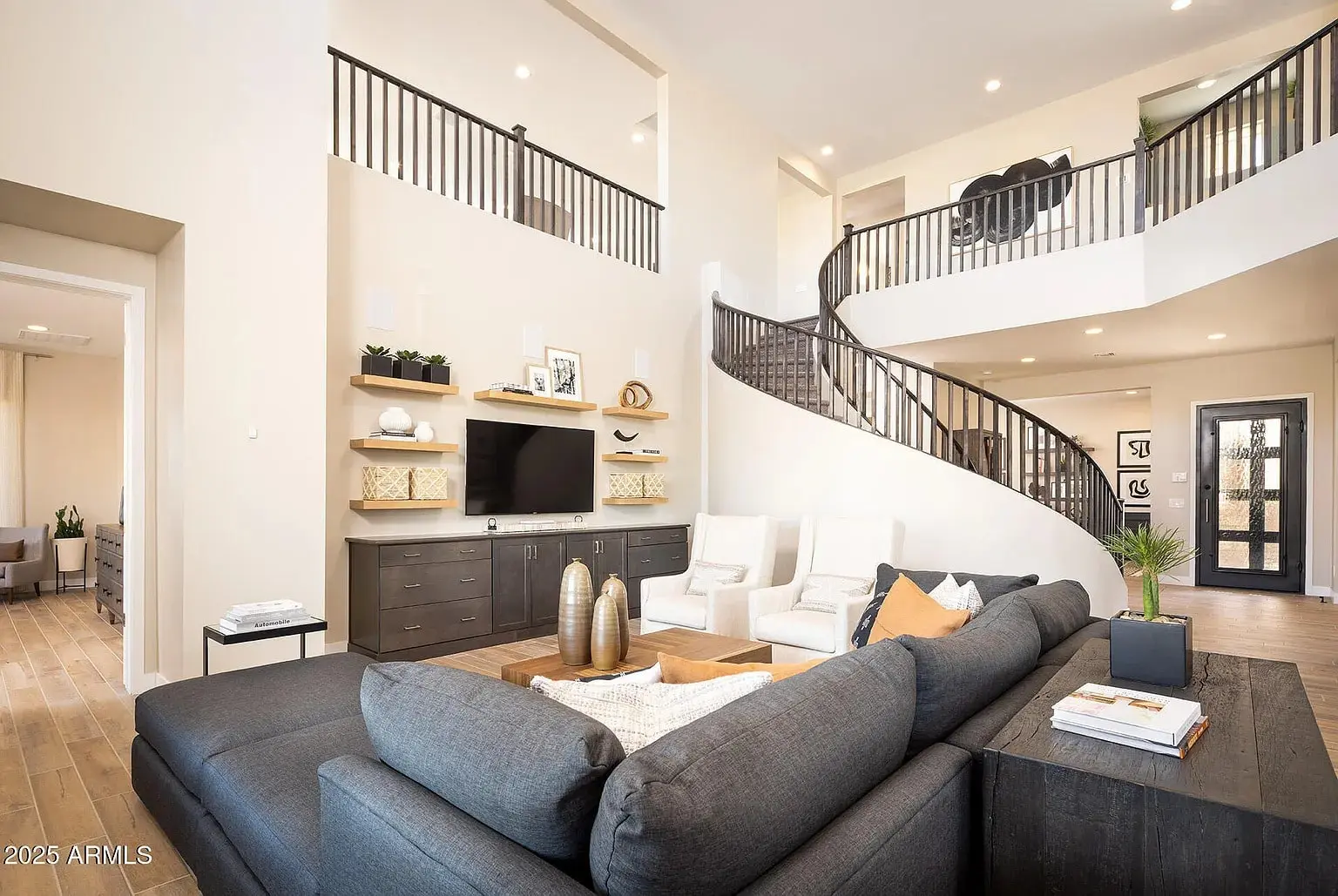 Modern living room with high ceilings, curved staircase, dark sectional sofa, and wall-mounted TV.