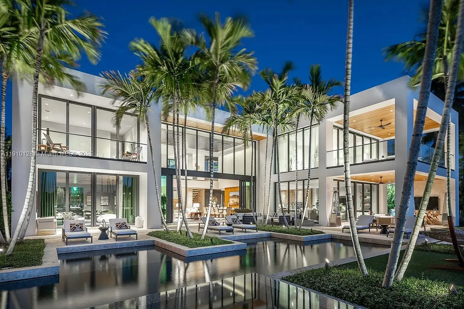 Modern luxury home with large glass windows, surrounded by palm trees and a reflecting pool.