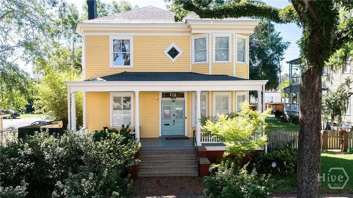 Yellow two-story house with a porch, bay window, and surrounding greenery.