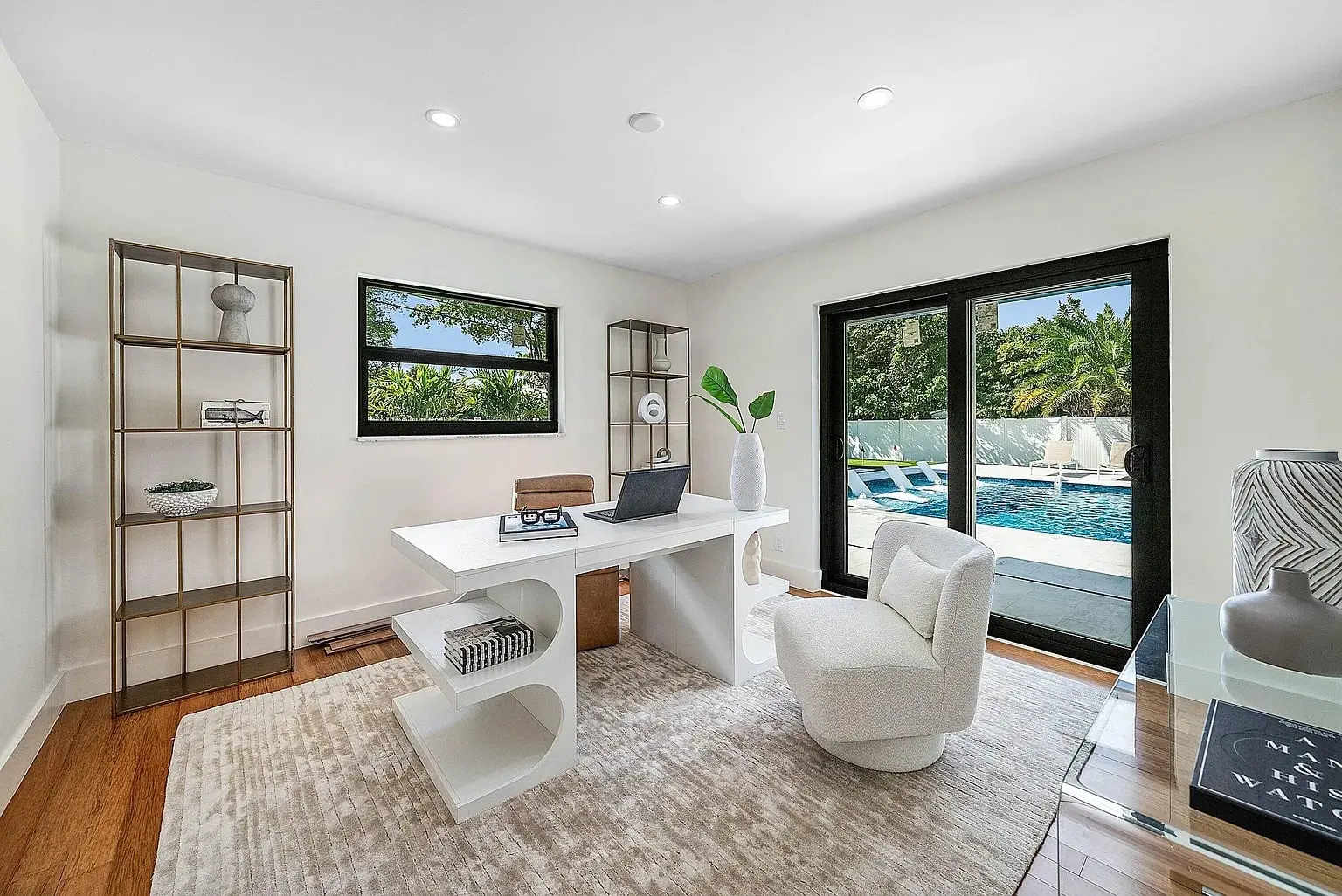 Modern home office with white desk, chair, and view of a pool through glass doors.