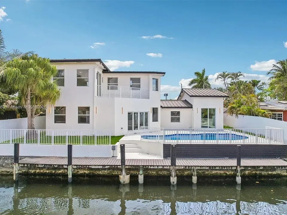 Modern white house with a pool, palm trees, and a dock by the water.