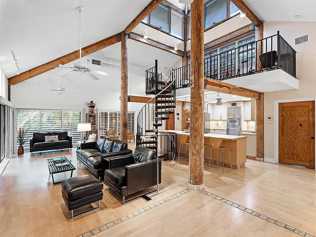 Spacious modern living room with high ceilings, wooden beams, black leather furniture, and a loft area.