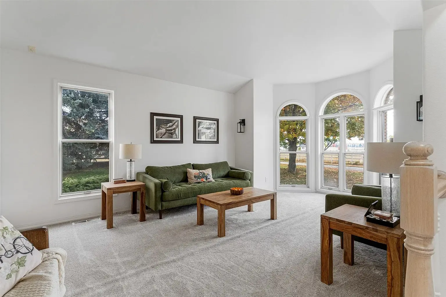 Bright living room with green sofas, wooden tables, and large arched windows overlooking trees.