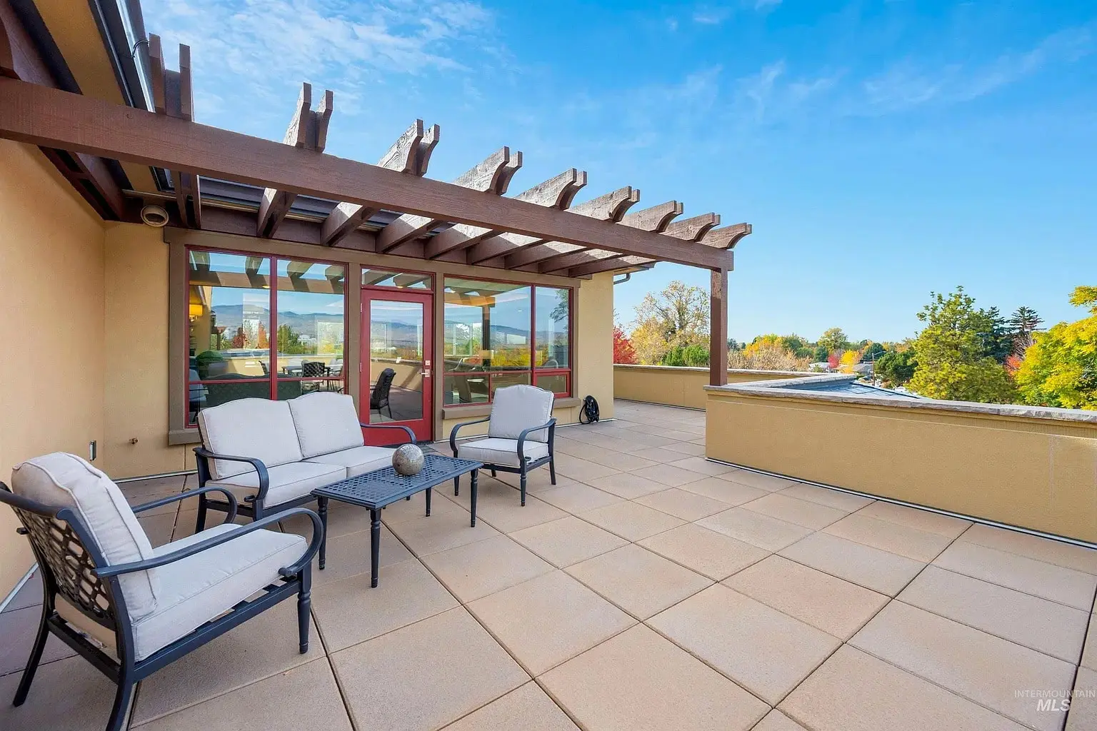 Spacious rooftop patio with cushioned seating, table, and pergola, overlooking trees and blue sky.