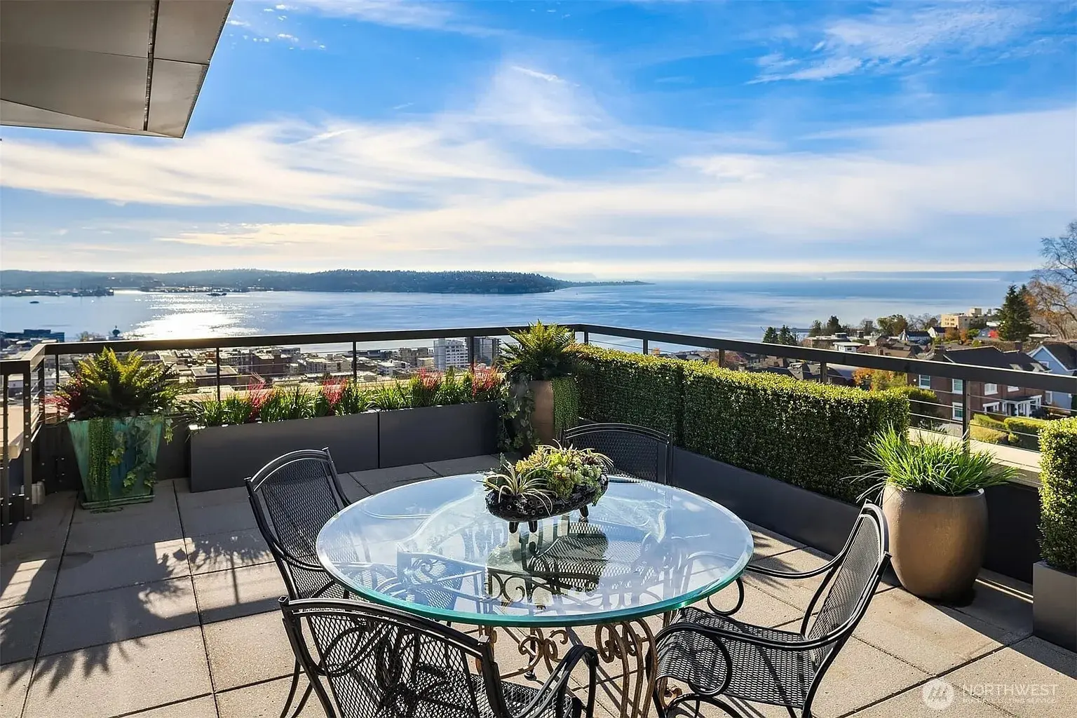 Rooftop patio with glass table, chairs, and ocean view under a clear blue sky.