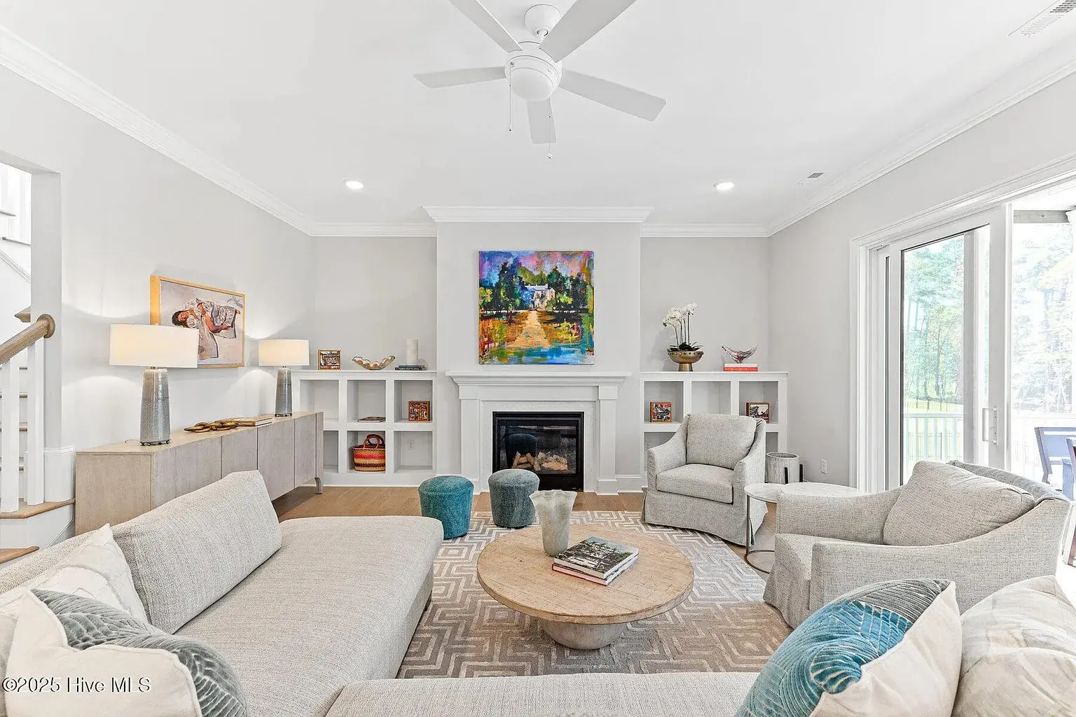 Modern living room with neutral tones, colorful artwork above fireplace, and large windows overlooking greenery.