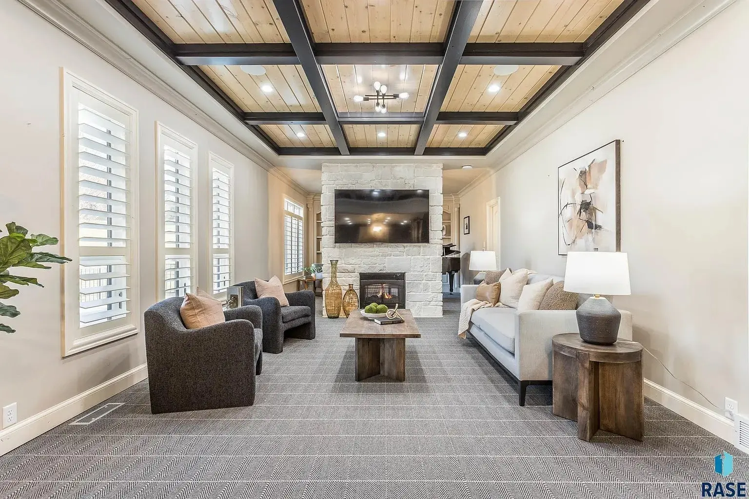 Modern living room with gray furniture, stone fireplace, large TV, and wooden ceiling beams.