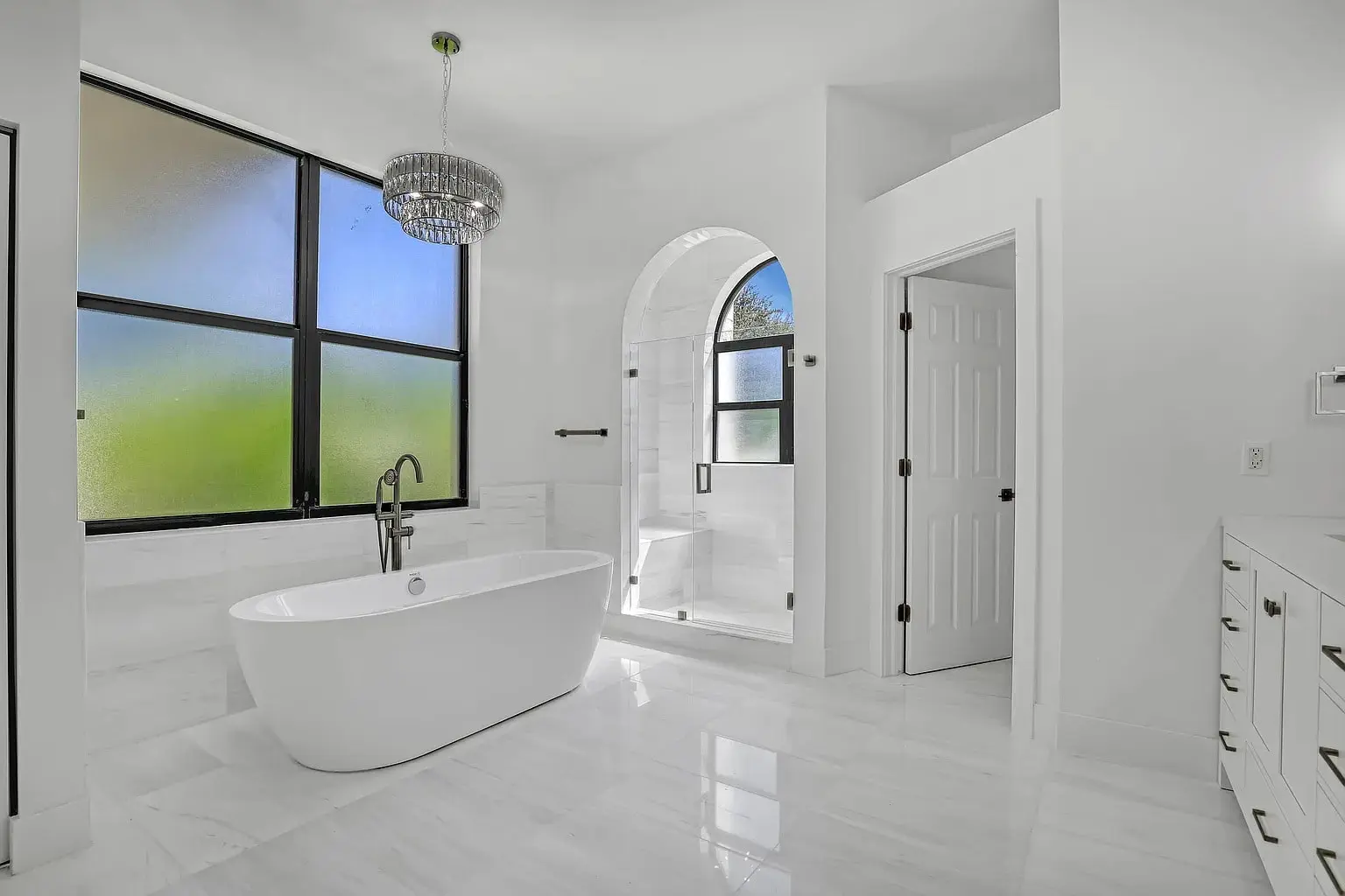 Modern bathroom with freestanding tub, glass shower, large frosted windows, and chandelier. White decor.