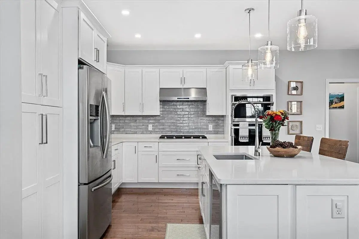 Modern white kitchen with stainless steel appliances, island, pendant lights, and a vase of flowers.