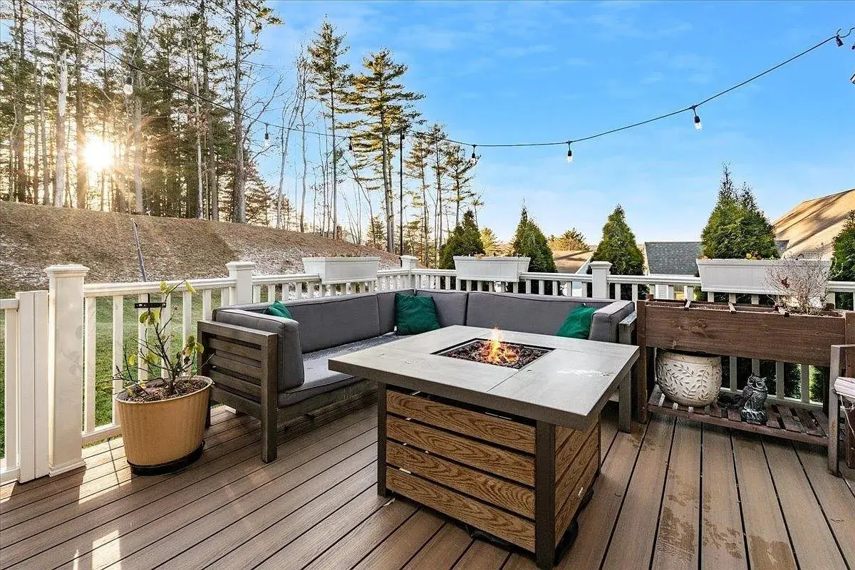 Outdoor deck with seating around a fire pit, surrounded by trees and string lights.
