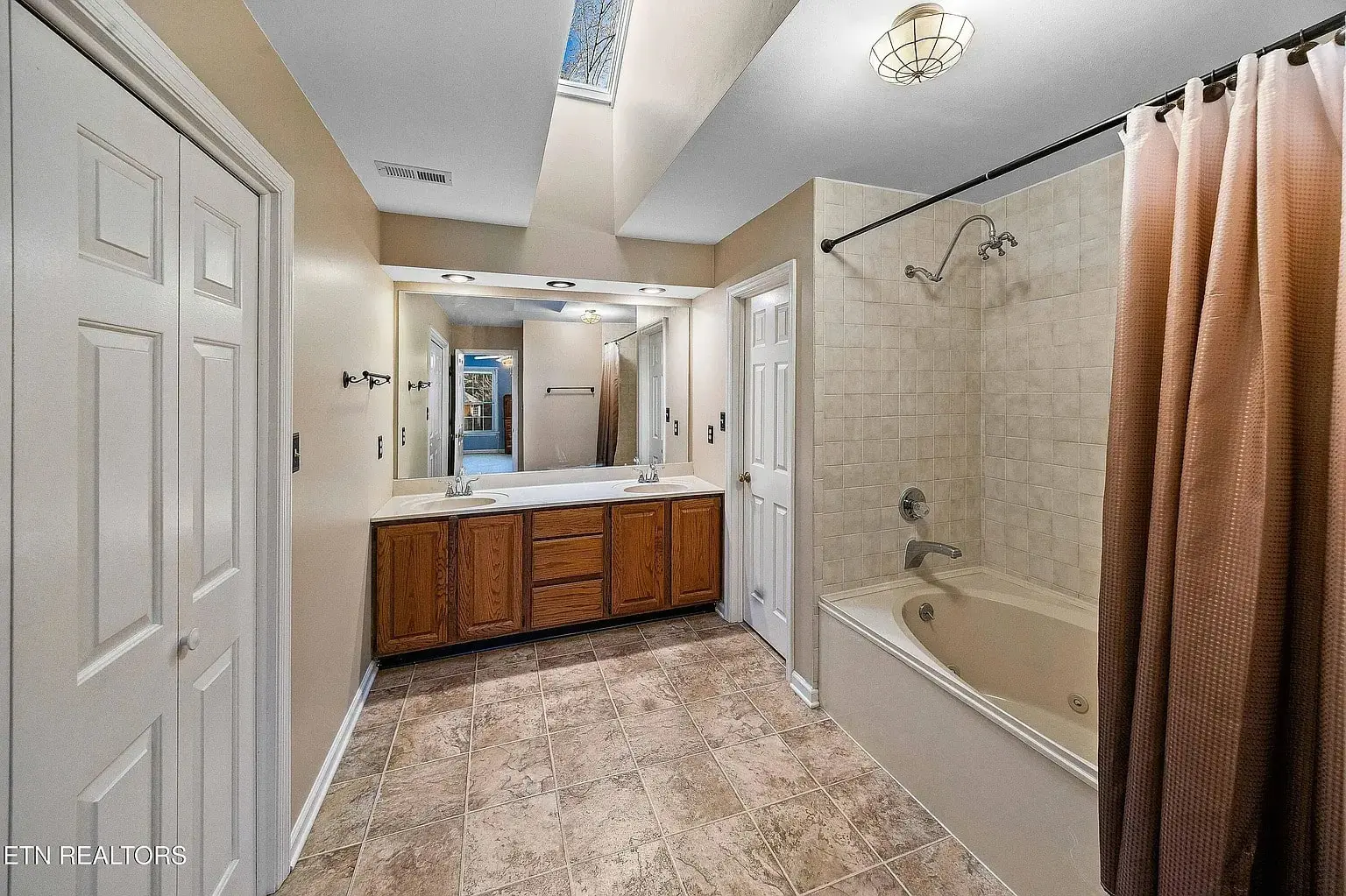 Spacious bathroom with double-sink vanity, skylight, tiled floor, and shower-tub combo with curtain.