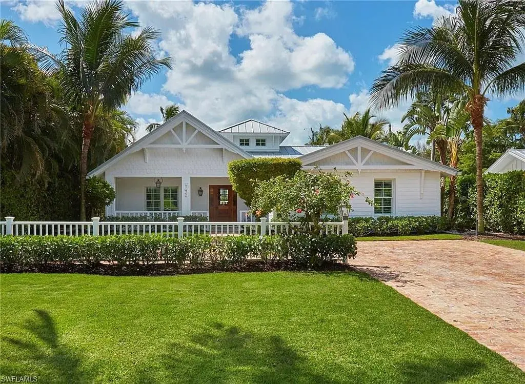 White single-story house with a brick driveway, surrounded by palm trees and a manicured lawn.