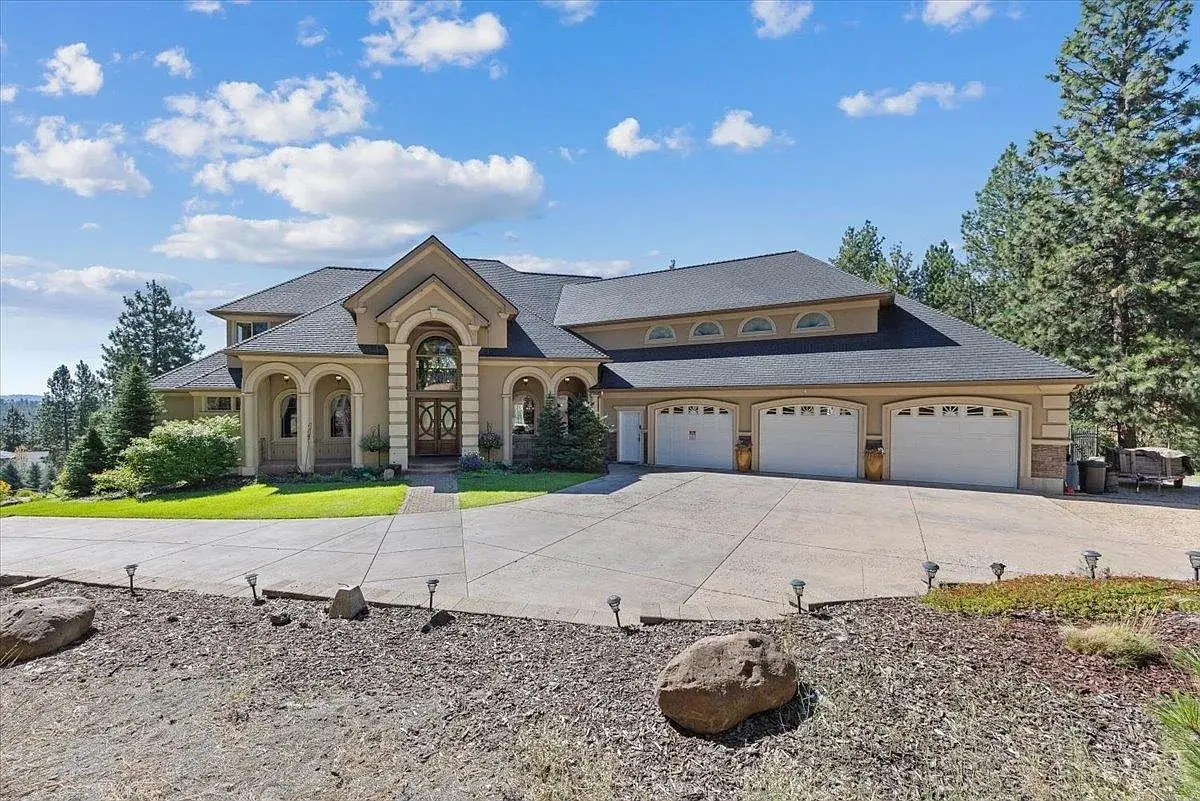 Large, elegant house with three-car garage, surrounded by trees and a clear blue sky.