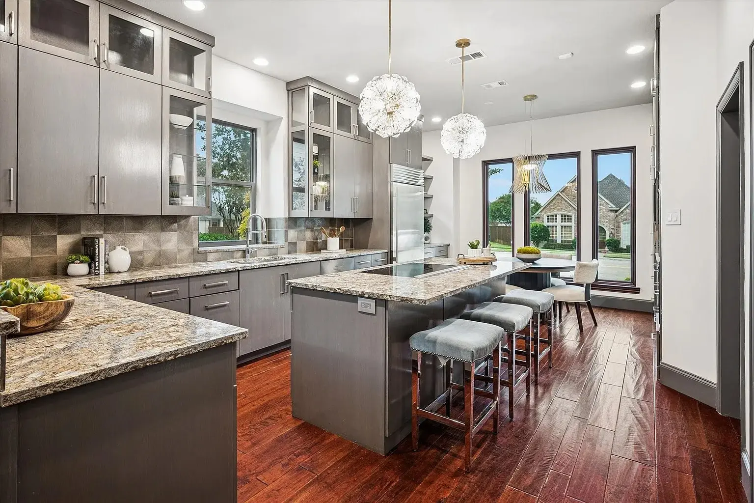 Modern kitchen with gray cabinets, granite countertops, island with barstools, and large windows overlooking a house.