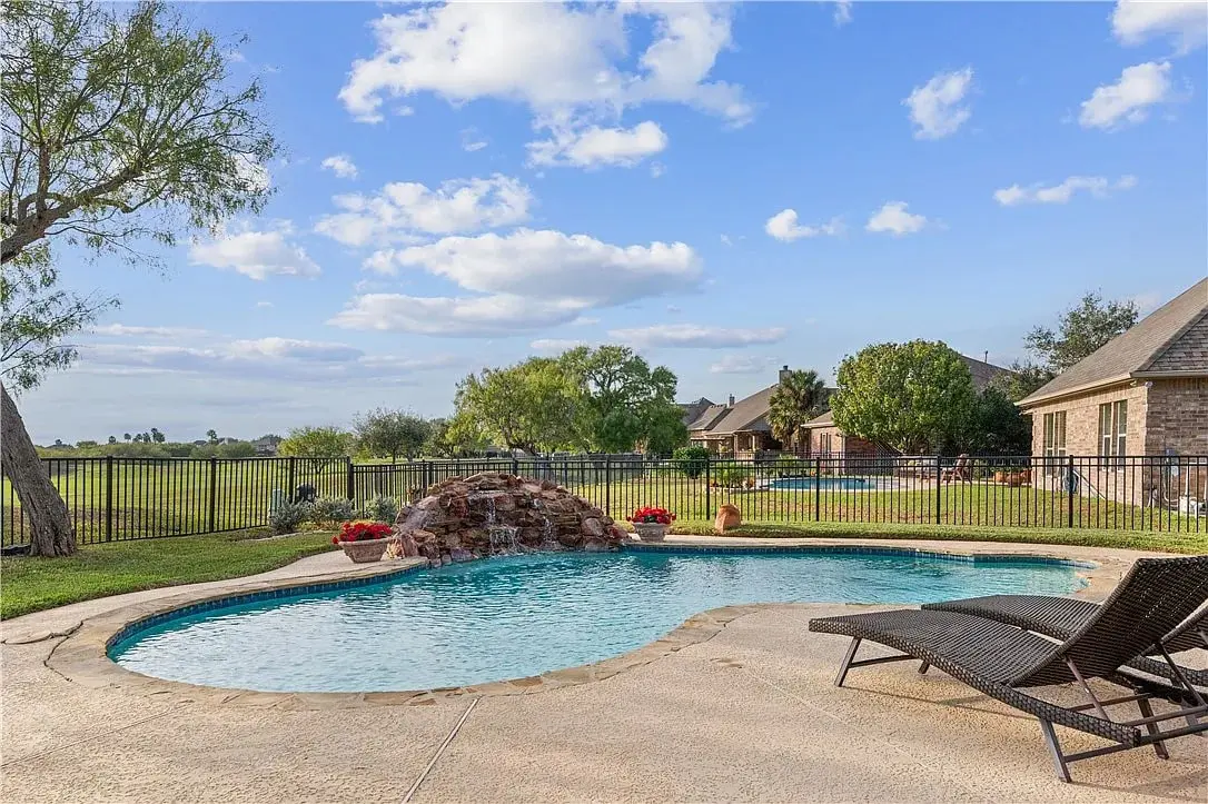 Backyard pool with waterfall feature, surrounded by trees and houses under a blue sky.
