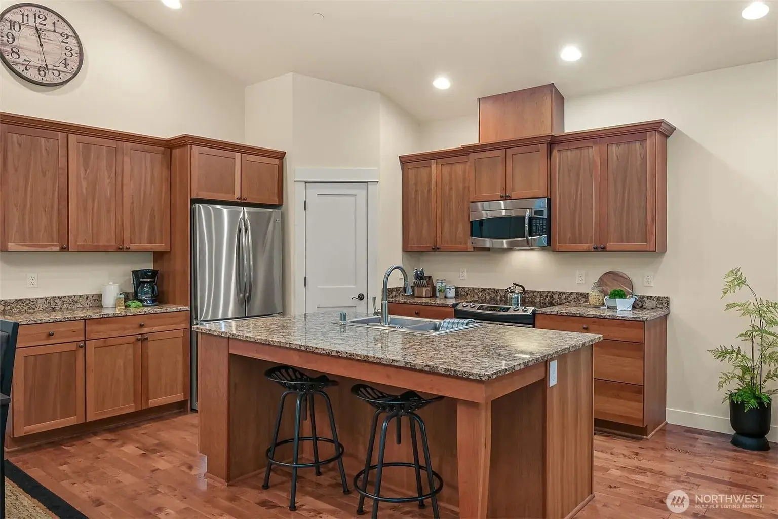 Modern kitchen with wooden cabinets, granite countertops, island with stools, stainless steel appliances, and clock.