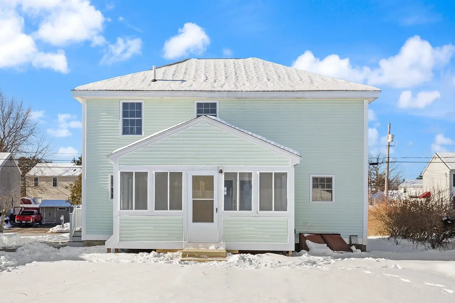 Two-story light green house with a white porch, surrounded by snow under a blue sky.