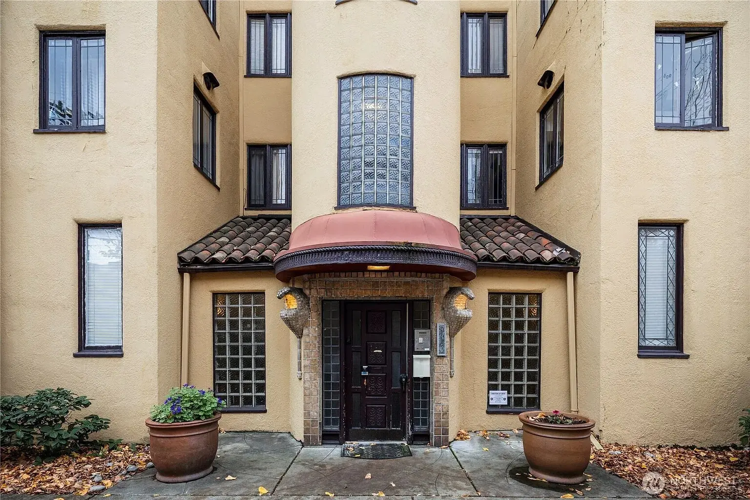Entrance of a beige building with a glass block window, brown door, and potted plants.