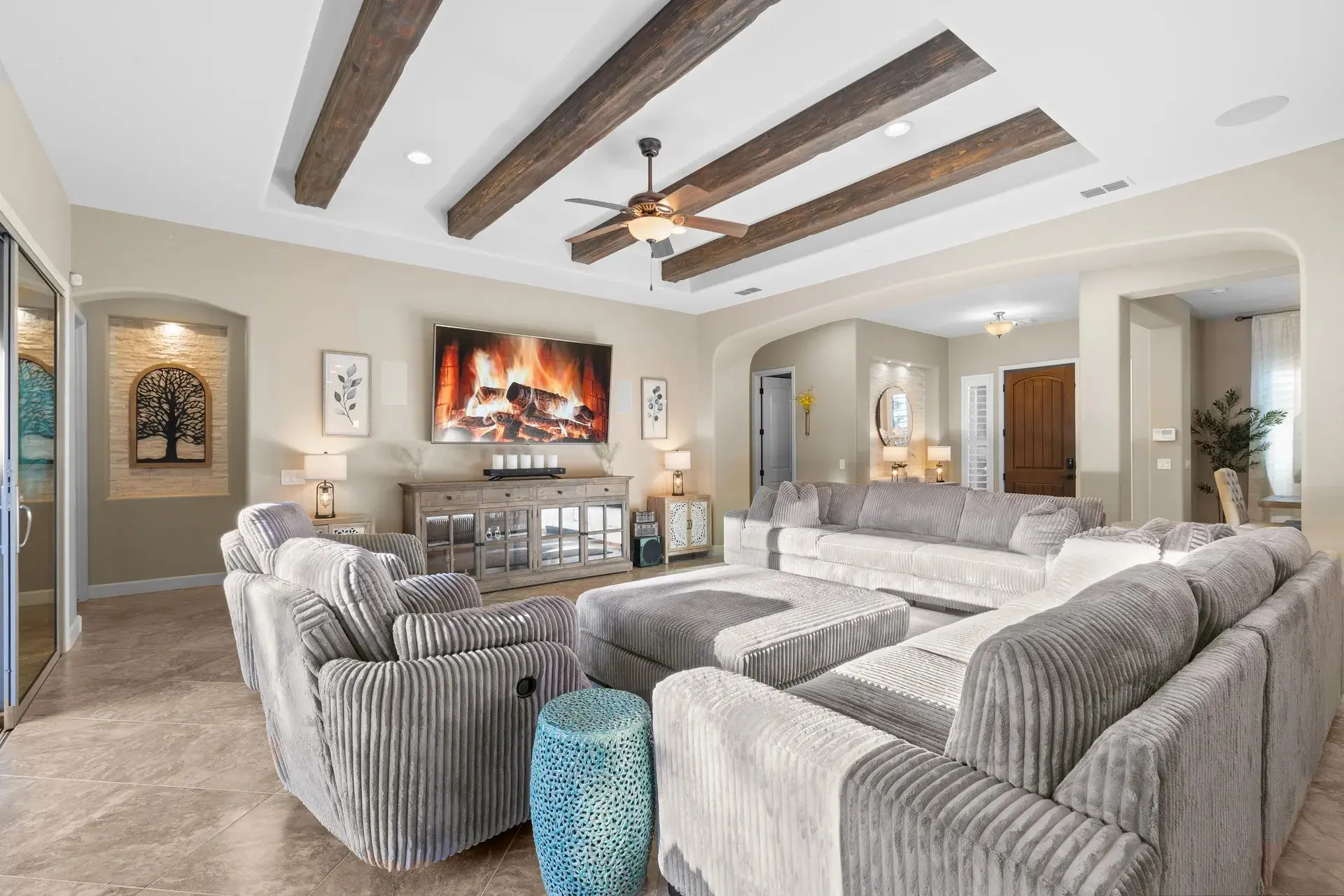 Bright living room with gray corduroy sectional, armchairs, TV fireplace, wooden beams, tile floor, sliding glass doors.